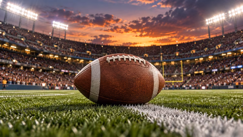 An American football rests on the turf beneath stadium lights at sunset before kickoff.